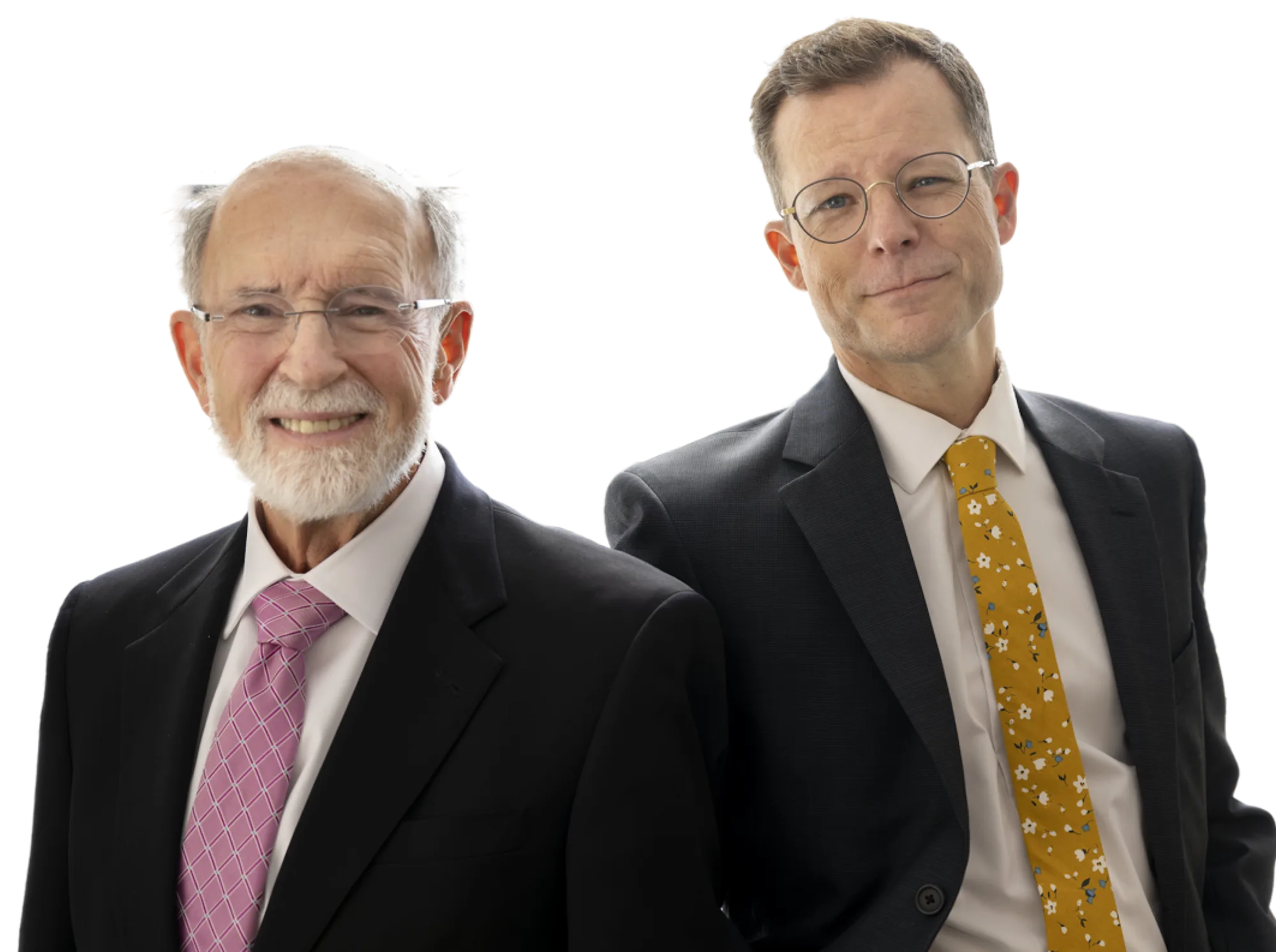 Two men in suits standing side by side, one older with white hair and beard wearing a pink tie, and one younger wearing glasses and a mustard yellow floral tie.