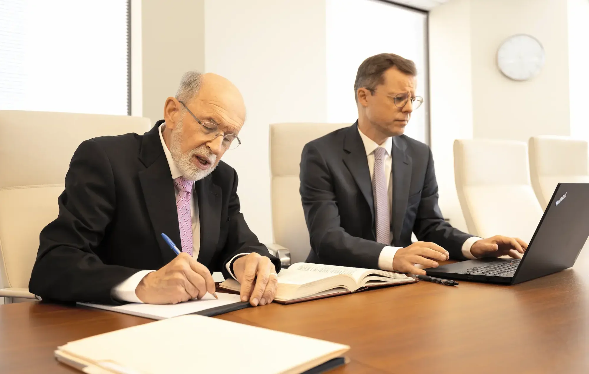 two men in suits sitting at a table with a laptop